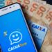 imagem mostra um celular com o aplicativo do Caixa Tem, em cima de um cartão e algumas notas de 100 e 50 reais.