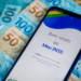 imagem mostra um celular com o aplicativo MEU INSS aberto com as boas vindas, e em baixo do celular algumas notas de dinheiro, notas de 100 e de 50 reais.