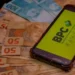 imagem mostra algumas notas de 100 e de 50 reais em baixo de um aparelho celular com a foto do BPC