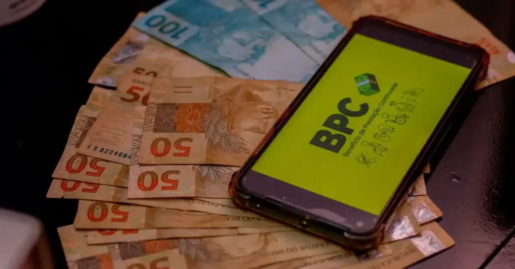 imagem mostra um celular com uma foto do BPC em cima de algumas cédulas de 100 e 50 reais